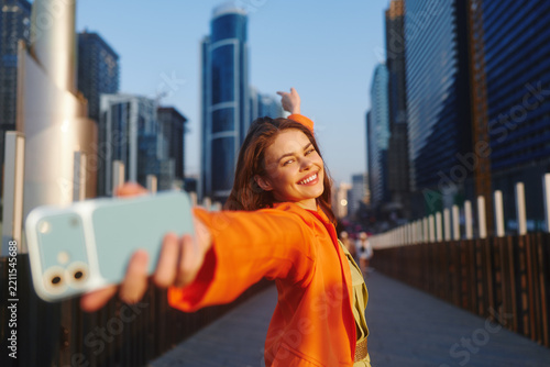 Smiling young woman in an orange jacket takes a selfie with a light blue smartphone, arm outstretched, against a city skyline of skyscrapers. Concept of mobile lifestyle and travel.
