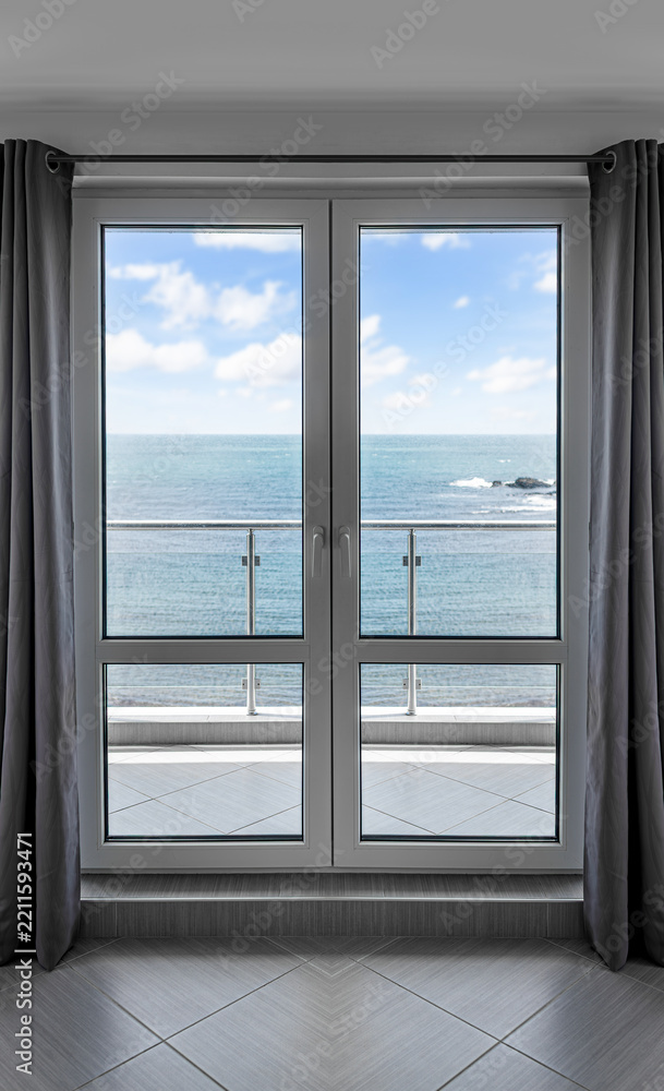 Fototapeta premium A modern glass balcony door with grey curtains frames a scenic view of the ocean under a blue sky.