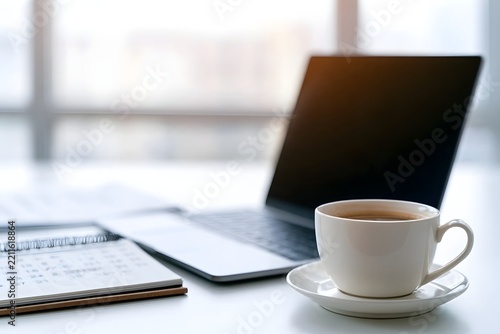 A white coffee cup sits beside a laptop and calendar on a bright desk  suggesting a work or study environment