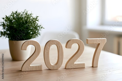 Wooden numbers 2027 arranged on a light wooden desk next to a small potted green plant in a neutral indoor setting