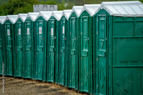 A row of green portable toilet cabins with white roofs stands on bare ground outdoors, with trees in the background.
