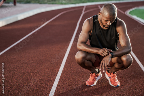 Young sportsman sitting on running track looking at smartwatch, fitness tracking, workout analysis, athlete recovery, sports technology, active lifestyle and outdoor training concept