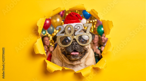 Funny pug wearing Santa hat and sparkly glasses with year numbers 2027, peeking through torn yellow paper background with party decorations. New Year celebration, festive pet, seasonal greeting.