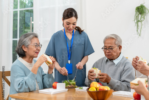 Female nurse accompanies senior Asian residents eating sandwiches during group dining in assisted living center, encouraging conversation, nutrition, companionship and active social engagement.