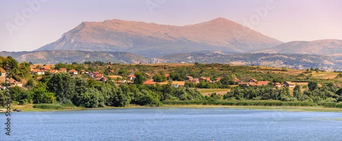Panel kuchenny z motywem Rural village with red roofs nestled among green trees and hills, overlooking a calm lake with Rtanj mountain in the background