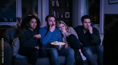 Friends on a couch in a dimly lit living room watching a scary movie and reacting with fear and surprise
