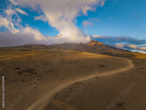 Aerial view of Volcano Chimborazo with a winding dirt road traversing the arid volcanic landscape under a clear blue sky during golden hour in Guano, Chimborazo, Ecuador.
