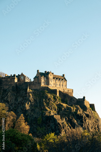 Daylight vertical photograph of the famous Edinburgh Castle perched on a rocky cliff in Scotland, surrounded by lush trees under a clear blue sky