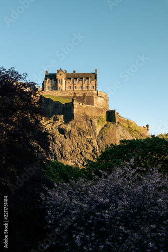 Daylight vertical photograph of the famous Edinburgh Castle perched on a rocky cliff in Scotland, surrounded by lush trees under a clear blue sky