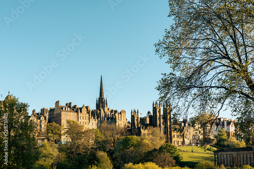 Daylight horizontal photograph showing the historic stone buildings and Gothic spire of Edinburgh's Old Town in Scotland, framed by trees under a clear blue sky