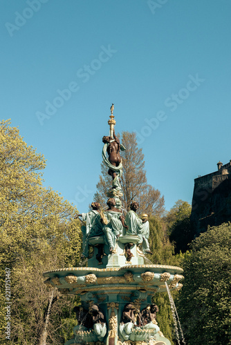 Vertical daylight photograph of the ornate Ross Fountain in Princes Street Gardens, Edinburgh, Scotland, featuring detailed turquoise and bronze statues against a clear blue sky