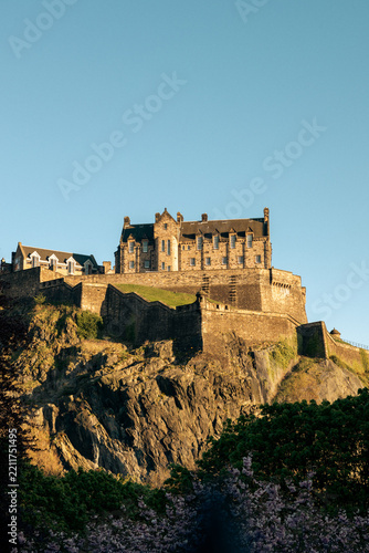 Daylight vertical photograph of the famous Edinburgh Castle perched on a rocky cliff in Scotland, surrounded by lush trees under a clear blue sky