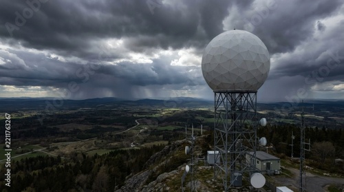 Large white weather radar station on a mountaintop against a stormy sky with distant rain over a scenic rural landscape.