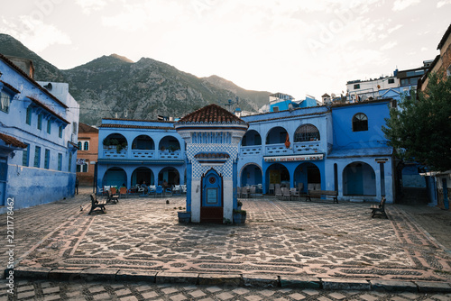 Wide daylight view of the lively cobblestone main square featuring traditional blue-painted arches, cafes, a central fountain pavilion, and the Rif mountains rising behind Chefchaouen
