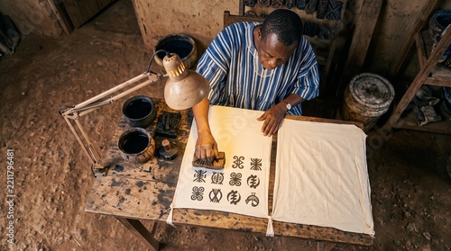 African artisan meticulously stamps traditional symbols onto fabric using wooden blocks and dark ink in a rustic workshop setting