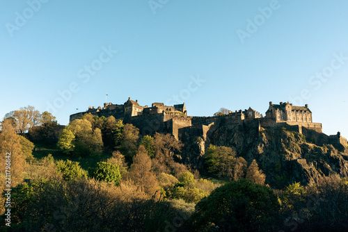 Wide horizontal daylight photograph of the iconic Edinburgh Castle perched on a rocky cliff in Scotland, surrounded by lush trees under a clear blue sky