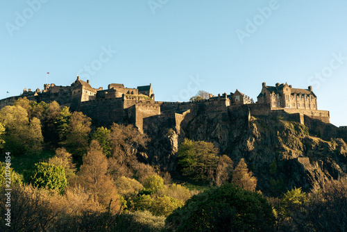 Wide horizontal daylight photograph of the iconic Edinburgh Castle perched on a rocky cliff in Scotland, surrounded by lush trees under a clear blue sky