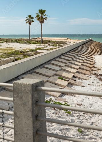  A concrete erosion control seawall structure stretches along the coast with two tropical palm trees under a bright blue sky at Fort De Soto Park, Florida.