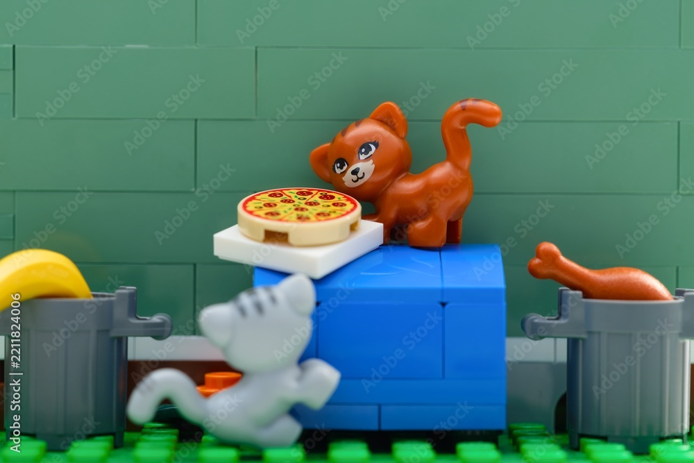 Obraz premium Tambov, Russian Federation - August 03, 2026 Lego ginger cat standing on a blue dumpster next to a pizza, while a grey cat and trash cans with food look on.