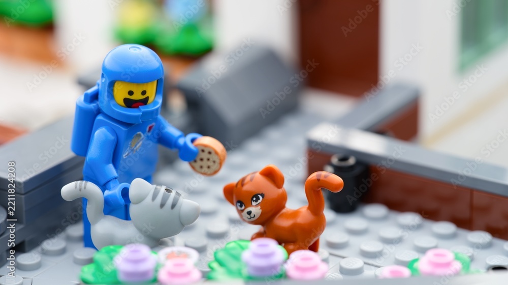 Obraz premium Tambov, Russian Federation - September 24, 2026 A blue Lego astronaut minifigure standing outside a house holding a miniature cookie, surrounded by a gray cat and a ginger cat.