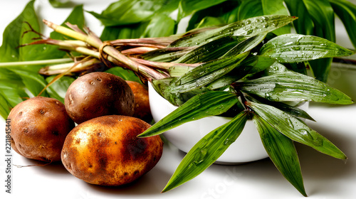 Fresh brown potatoes and bundle of wet green leaves in white bowl on white background, close up.