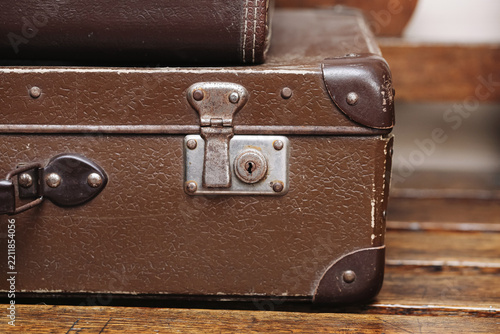 Vintage brown suitcase with metal lock and worn corners on wooden floor. Old travel luggage with textured surface and leather handle. Retro baggage background. Aged case and rusty hardware.