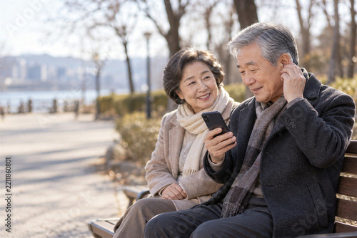 Senior asian man adjusting hearing aid while wife shows smartphone app on park bench, hearing aid technology