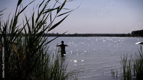 A silhouetted person stands in sparkling lake water with arms raised. Tall green reeds frame the foreground under bright natural sunlight across the calm surface.