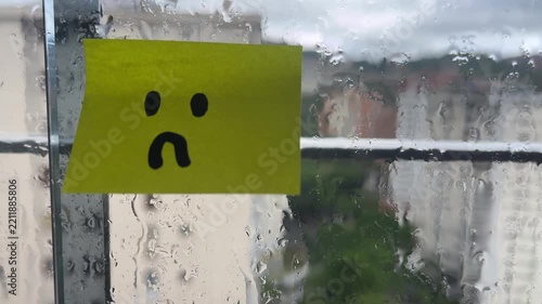 Sad face sticky note on rainy window stuck indoors