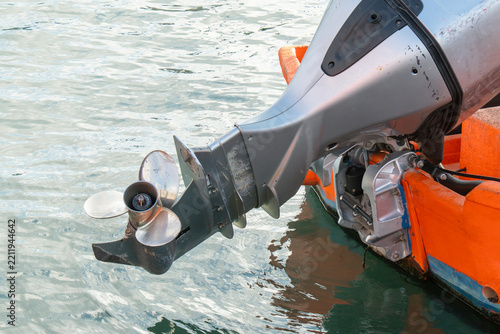 Close-up of a metallic outboard motor mounted on an orange wooden boat floating on calm water, showing the propeller and engine details.