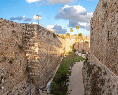 Historic stone fortress walls surround a peaceful pathway in Acre, Israel, with palm trees, dramatic clouds, and blue sky.