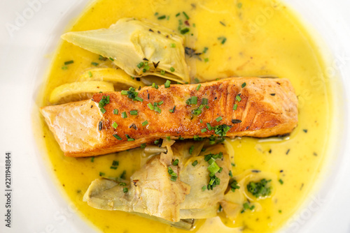 Seared salmon fillet served with artichoke hearts and fresh herbs in a creamy yellow sauce on a white plate.