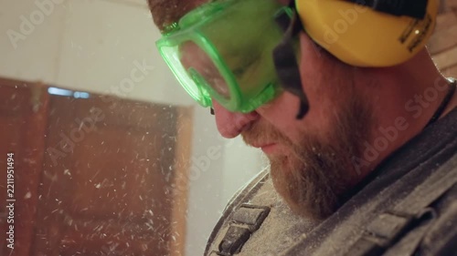 Close up shows part of man face wearing green safety glasses and yellow ear protection with wood dust particles visible in air. scene is indoors likely during woodworking or carpentry
