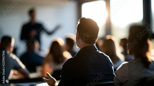 Adult education teacher in front of his class view from the back of the classroom toward a teacher in a soft focus silhouette against a whiteboard adult students seated at desks in the