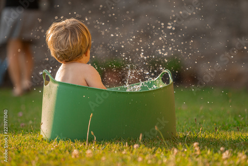 A young child sits in a green outdoor tub splashing water on a sunny lawn, with droplets catching the light and a dog visible in the background.