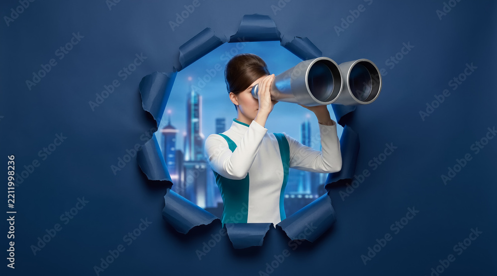 Fototapeta premium Woman looking through binoculars via a hole in blue paper, night cityscape visible behind, concept of vision and discovery. Business, exploration, future planning.