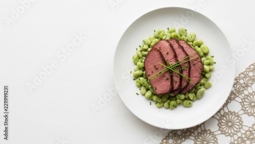Sliced smoked beef Judd mat Gaardebounen with creamy broad beans and chives on a stark white linen background, featuring a delicately embossed muted gold foil crochet lace motif and left copy space