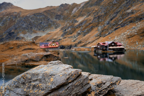 Scenic autumn landscape at Balea Lake in the Fagaras Mountains, Romania, featuring textured rocks in the foreground and a mountain cabin reflected in the calm water.