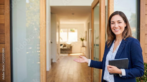 Smiling real estate professional stands at the open front door of a modern house and gestures for visitors to enter the bright residence while
