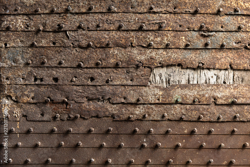 Close-up of a weathered rusty metal door with rows of rivets and visible corrosion, revealing some underlying wood through damaged sections