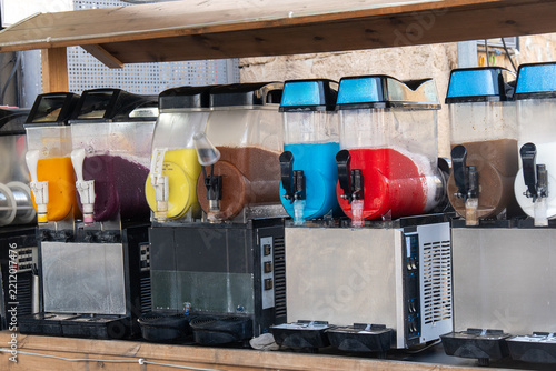 Multiple slush machines with various colorful frozen drinks are displayed on a wooden counter in an outdoor setting, ready for serving.