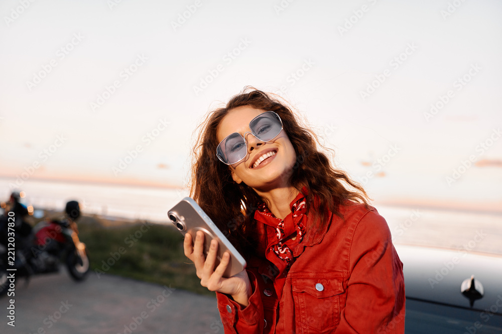 Obraz premium Young woman in a red jacket smiles while looking at a smartphone outdoors at sunset, with a calm water backdrop. Candid lifestyle shot for social, travel, or fashion use. Great for branding.