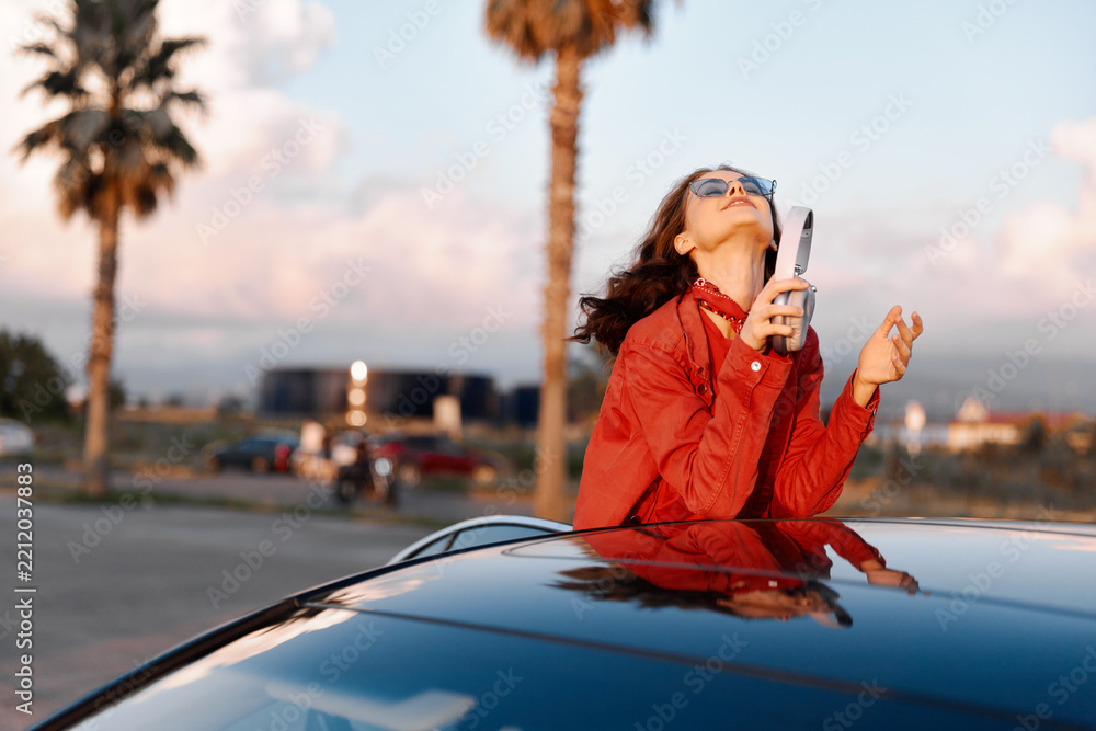 Obraz premium Young woman in red jacket and sunglasses leaning on car roof, holding smartphone under palm trees. Carefree travel and summer vacation mood at sunset.