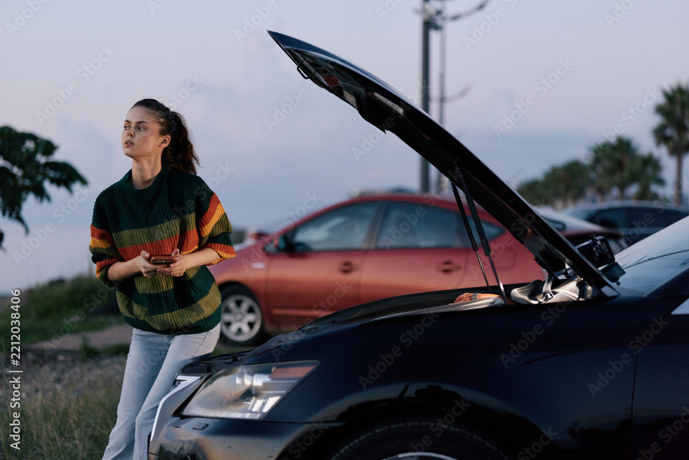 Obraz premium Young woman in a striped sweater stands beside a car with the hood up, glancing at the engine with worry. Outdoor roadside scene, casual travel trouble, ready to troubleshoot.