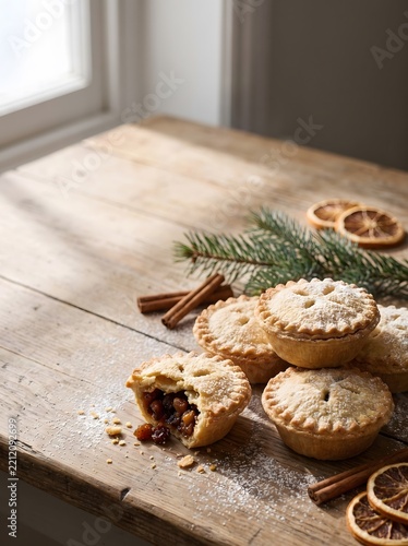 Close-up photo of festive mince pies dusted with sugar on a rustic wooden table beside cinnamon sticks, dried orange slices and a fir branch. Warm natural light food photography, holiday