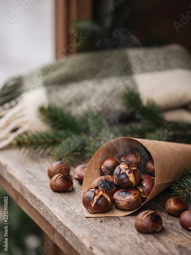 Roasted chestnuts in a rustic wooden bowl surrounded by fresh chestnuts and evergreen branches. Warm photorealistic food photography with cozy seasonal styling and natural textures, ideal for autumn
