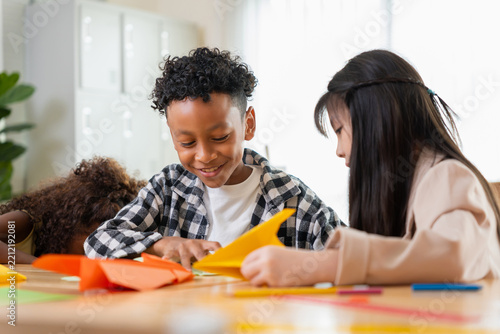 Multiethnic kids doing origami paper craft in classroom, diverse school children folding paper in kindergarten class, cute african american boy and asian girl learning arts and crafts at school desk