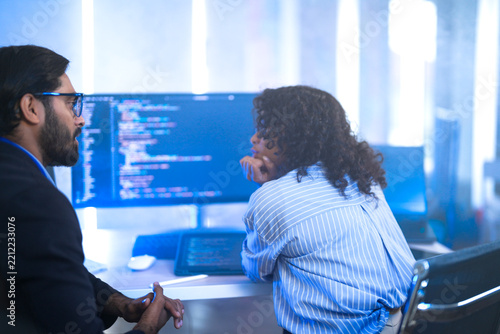 A male team lead leaning over a seated female developer explaining a fix on the monitor, to point at code on her monitor in a blue-lit office; over-the-shoulder angle of software frame6489.