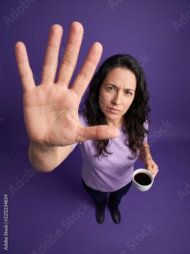 Serious Woman Holding Coffee With Hand Raised Stop Gesture Against Purple Background

