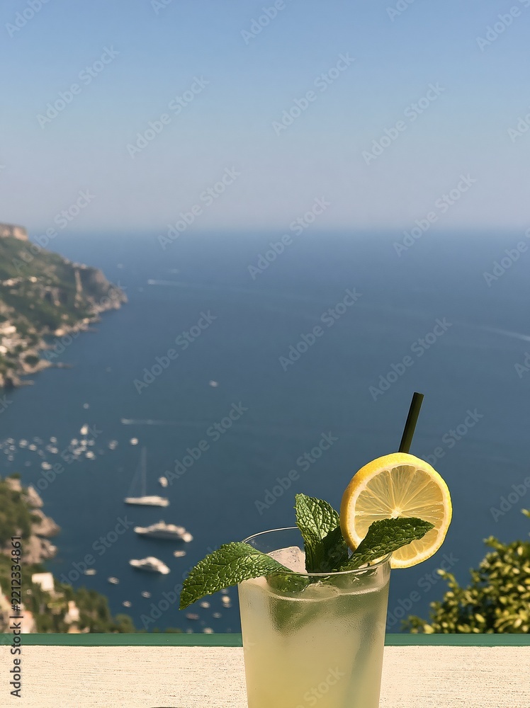 Fototapeta premium Refreshing Lemonade Cocktail with Mint and Lemon Slice Overlooking Scenic Coastal Town and Ocean View